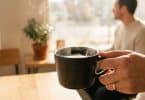 Une main tient une tasse de café fumante dans un café lumineux avec une personne en arrière-plan.