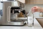 Une main tenant un petit verre doseur au-dessus d'une bouteille de vinaigre blanc à côté d'une machine à café.