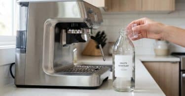Une main tenant un petit verre doseur au-dessus d'une bouteille de vinaigre blanc à côté d'une machine à café.