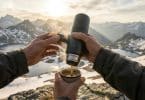 Mains utilisant une cafetière portable pour servir un café chaud face à un paysage montagneux enneigé au coucher du soleil.