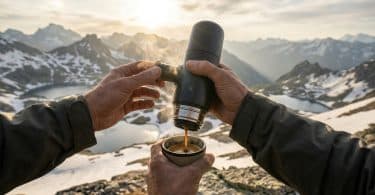 Mains utilisant une cafetière portable pour servir un café chaud face à un paysage montagneux enneigé au coucher du soleil.