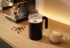 Un mousseur à lait noir électrique en action avec du lait chaud, à côté d'une tasse de cappuccino et d'une machine à café.