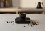 Une petite tasse à expresso en céramique posée sur une balance électronique noire, entourée de grains de café.