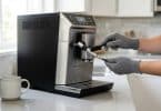 Une personne portant des gants gris nettoie la buse d'une machine à café avec une brosse dans une cuisine lumineuse.