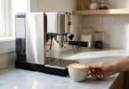 Une personne tient une tasse de cappuccino devant une machine à café espresso en acier inoxydable dans une cuisine.