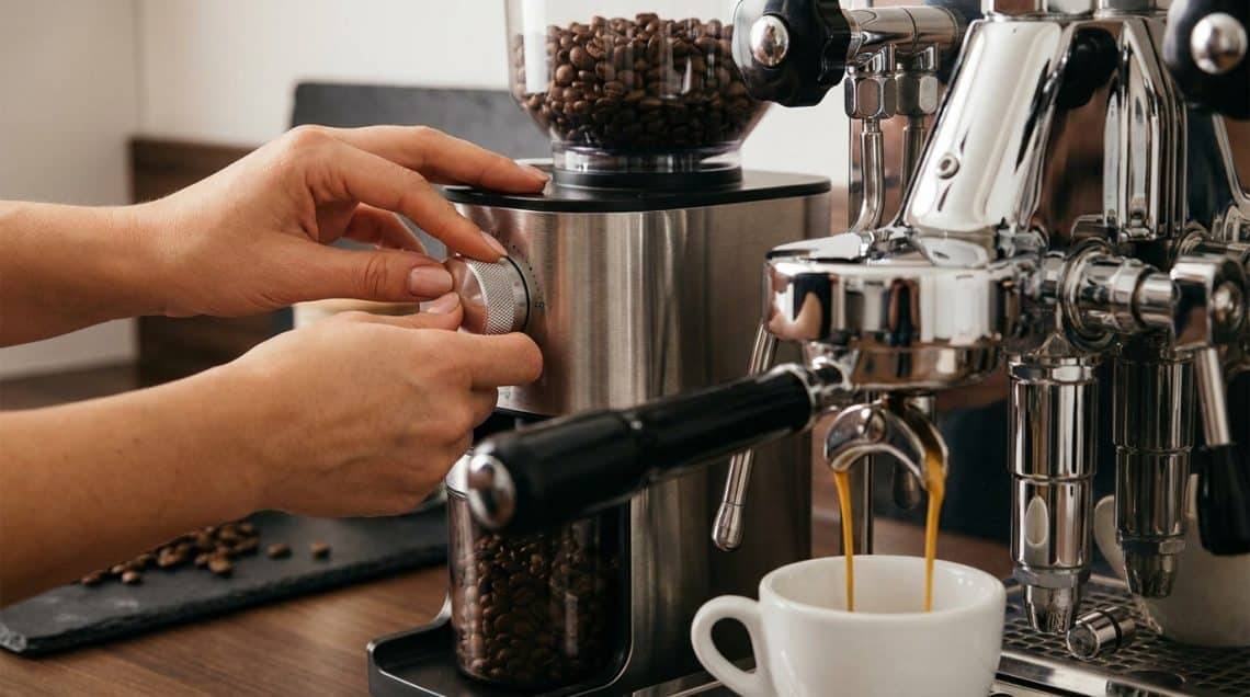 Mains réglant la finesse de mouture sur un broyeur à café inox, à côté d'une machine expresso professionnelle.