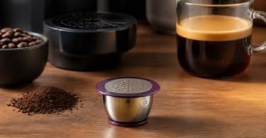 Capsule de café réutilisable en inox avec bord violet posée sur une table en bois à côté d'une tasse de café.