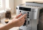 Une main règle la finesse de mouture sur une machine à café DeLonghi Magnifica S dans une cuisine lumineuse.