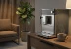 Machine à café moderne en inox posée sur un meuble en bois dans une salle d'attente élégante avec une plante verte.