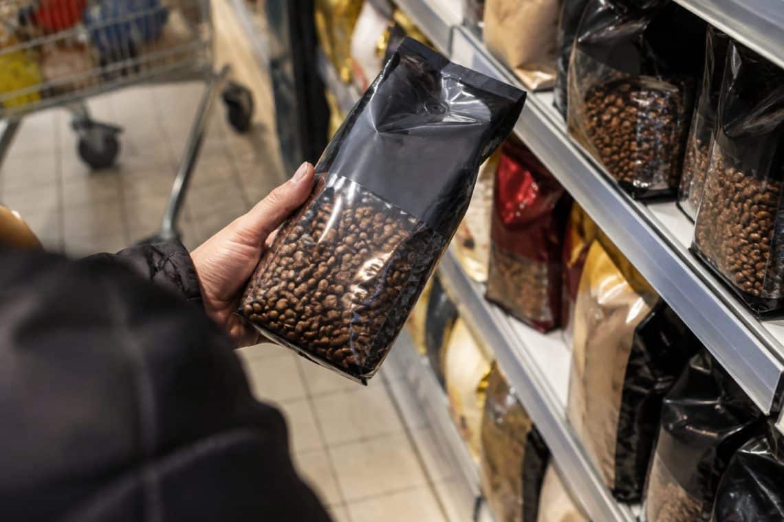 Une main tient un sachet de caf&eacute; en grains noir devant le rayon d'un supermarch&eacute;.