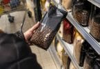 Une main tient un sachet de café en grains noir devant le rayon d'un supermarché.