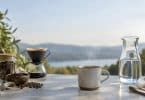 Une tasse de café fumant, une carafe d'eau et du matériel de préparation sur une table face à un lac.