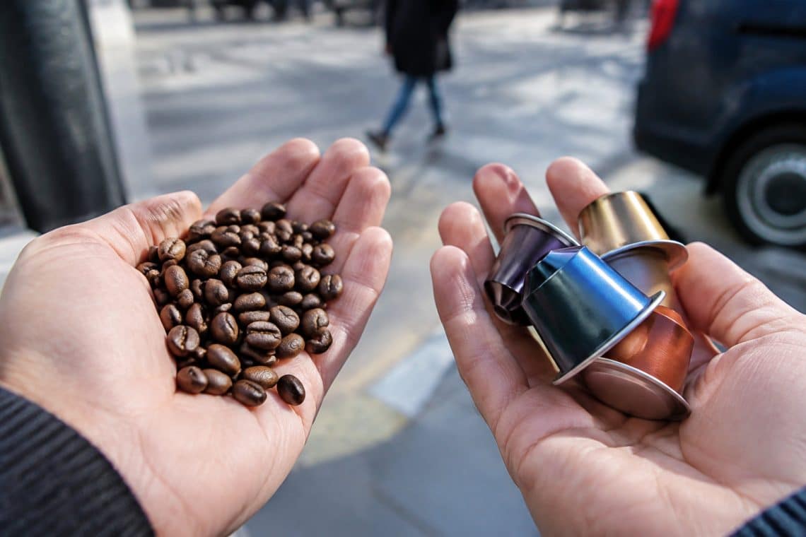 Mains tenant des grains de caf&eacute; d'un c&ocirc;t&eacute; et des capsules color&eacute;es de l'autre, avec une rue floue en arri&egrave;re-plan.