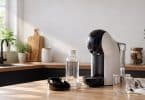 Machine à café Dolce Gusto Genio S sur un plan de travail avec une bouteille de solution de détartrage et un verre vide.