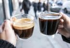 Deux mains tiennent chacune une tasse en verre contenant un café, l'un avec une mousse épaisse et l'autre noir profond.
